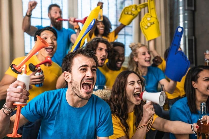 Fãs esportivos multinacionais e alegres assistindo ao jogo na TV em casa.