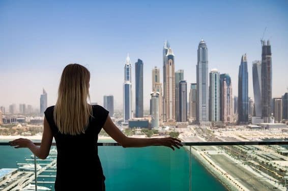 Uma mulher de preto com expressão pensativa em uma sacada com vista para os arranha-céus de Dubai, Emirados Árabes Unidos.