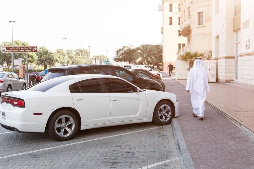 Um homem em trajes tradicionais e um Dodge branco em um estacionamento.