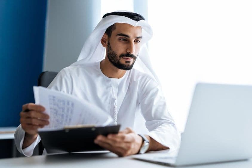 Homem trabalhando com laptop em escritório em Dubai, vestindo traje tradicional branco Emirati.
