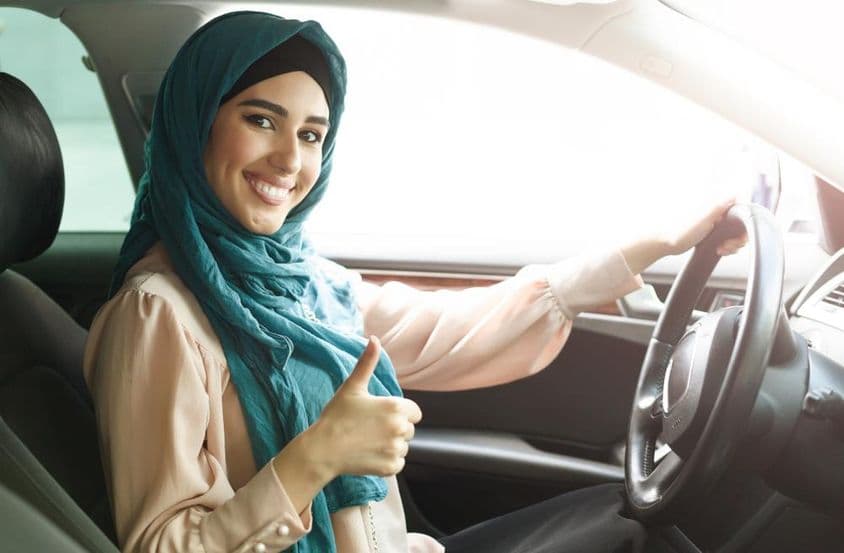 Mulher árabe feliz de hijab dirigindo seu carro com um sinal de positivo.