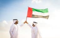 Homens árabes vestindo kanduras caminhando no deserto segurando uma bandeira.