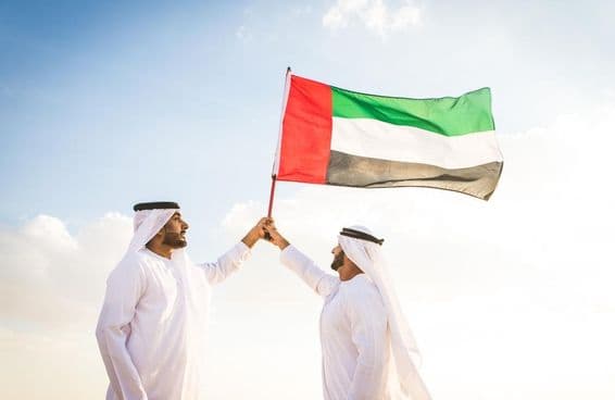 Homens árabes vestindo kanduras caminhando no deserto segurando uma bandeira.