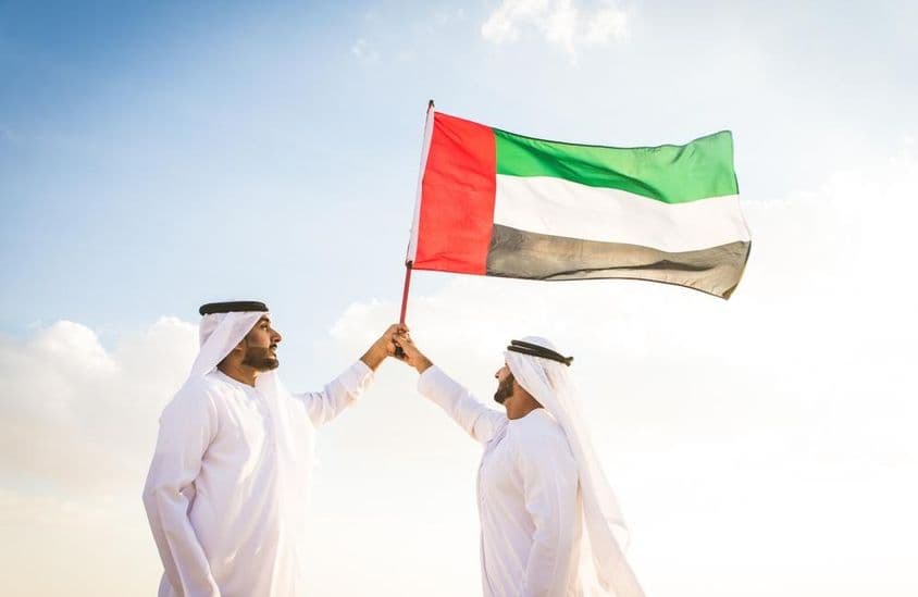 Homens árabes vestindo kanduras caminhando no deserto segurando uma bandeira.