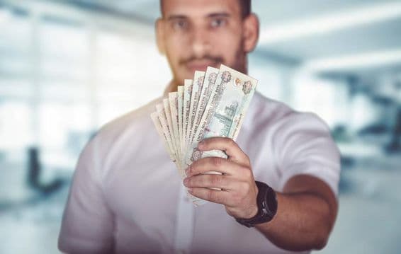 Homem segurando cédulas de dirham, em frente a uma agência bancária.