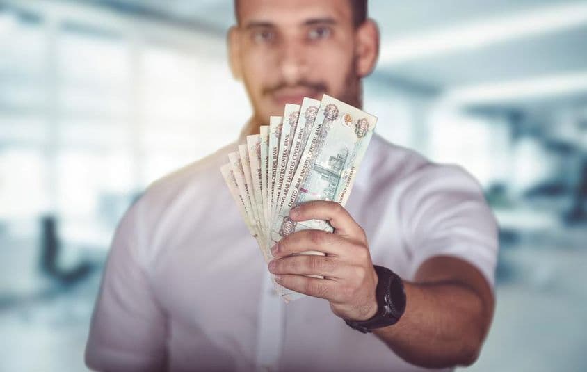 Homem segurando cédulas de dirham, em frente a uma agência bancária.