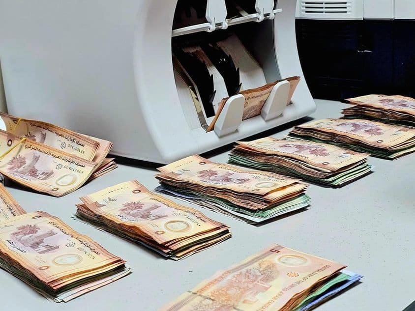 Máquina de contar dinheiro com maços de notas de dirham emiradense organizados em uma mesa.