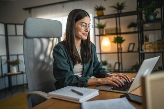Mulher adulta trabalhando em um laptop e usando um caderno em um escritório doméstico.