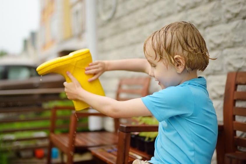 Menino de pré-escola enxarcado despejando água de uma bota de borracha amarela.