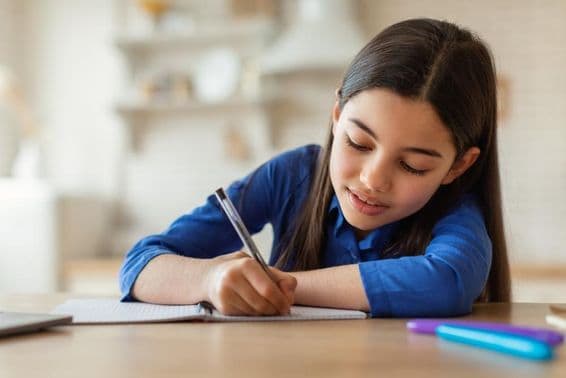 Menina árabe tomando notas enquanto está sentada à mesa com um laptop em casa.