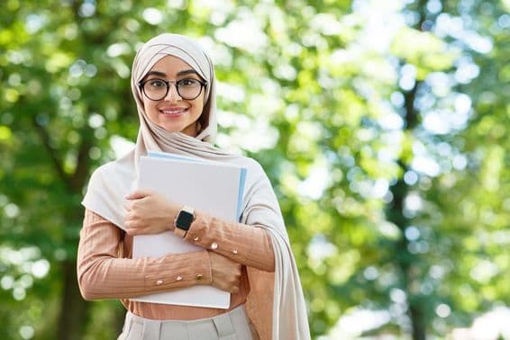 De volta às aulas, mulher árabe de hijab e óculos segurando livros em parque.