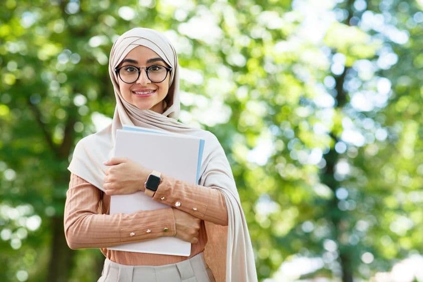 De volta às aulas, mulher árabe de hijab e óculos segurando livros em parque.