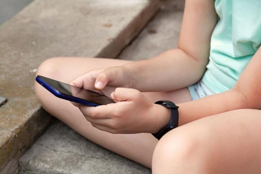 Uma criança está sentada ao ar livre usando um smartphone.