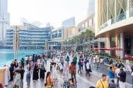 Uma multidão diversificada de turistas e locais caminha ao longo da movimentada promenade à beira-mar em frente ao Dubai Mall.
