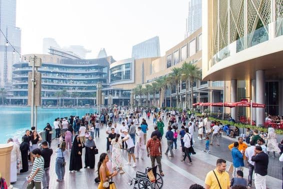 Uma multidão diversificada de turistas e locais caminha ao longo da movimentada promenade à beira-mar em frente ao Dubai Mall.